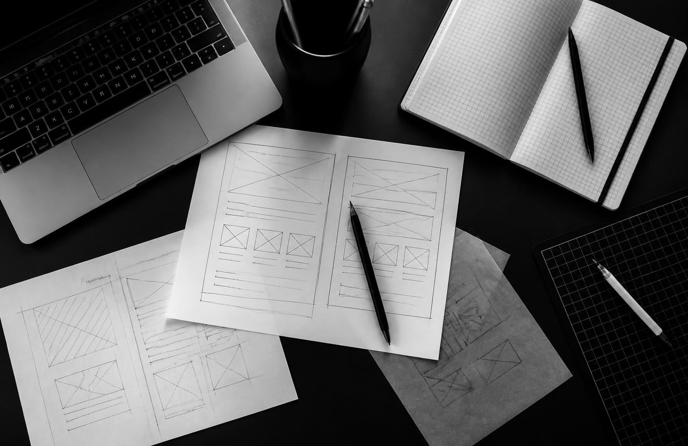 Overhead view of interface wireframe sketches, a notebook and a laptop on a dark desk